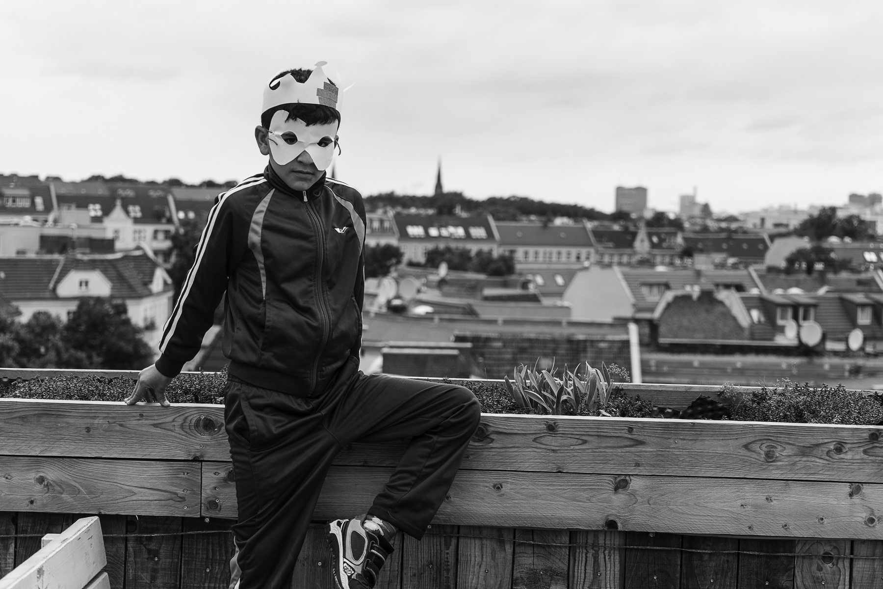 Ein Junge mit einer Papierkrone und einer Maske steht auf einem Dach und blickt auf die Stadt hinunter. (KI generierte Beschreibung)