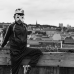 Eine Person in einem Trainingsanzug und einer Tiermaske lehnt an einem Holzgeländer, im Hintergrund sind eine Stadtlandschaft und ein bewölkter Himmel zu sehen. (KI generierte Beschreibung)