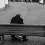 Ein älterer Mensch sitzt allein auf einer Holzbank in einem leeren, gepflasterten Außenbereich und wendet der Kamera den Rücken zu. (KI generierte Beschreibung)