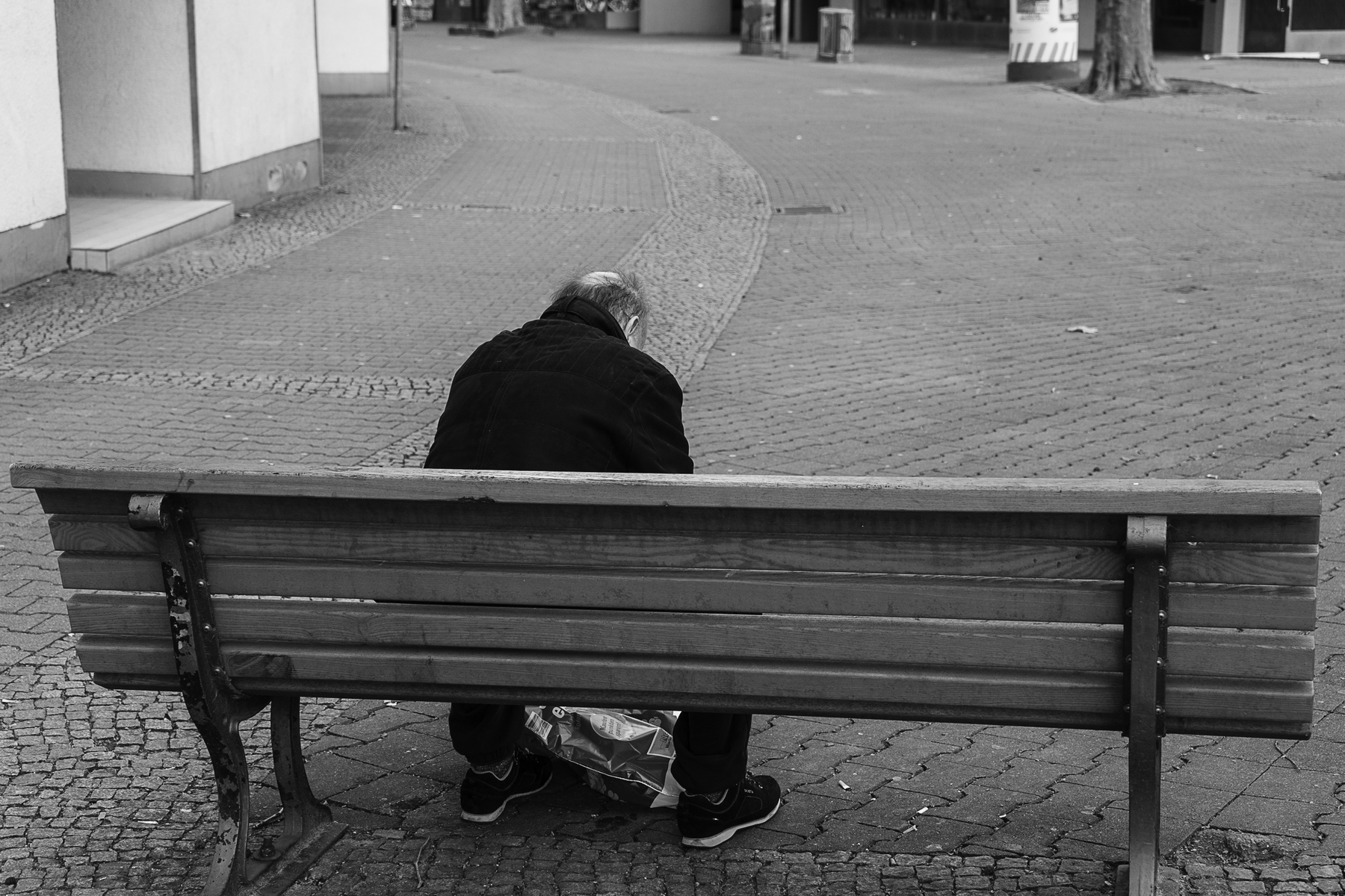 Ein älterer Mensch sitzt allein auf einer Holzbank in einem leeren, gepflasterten Außenbereich und wendet der Kamera den Rücken zu. (KI generierte Beschreibung)