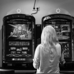 Eine Person mit hellem Haar steht in einem schwach beleuchteten Raum vor zwei Spielautomaten. (KI generierte Beschreibung)