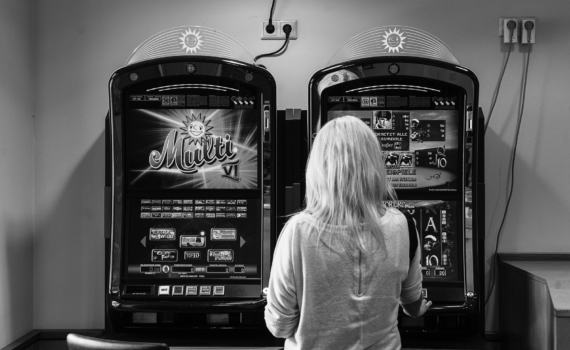 Eine Person mit hellem Haar steht in einem schwach beleuchteten Raum vor zwei Spielautomaten. (KI generierte Beschreibung)
