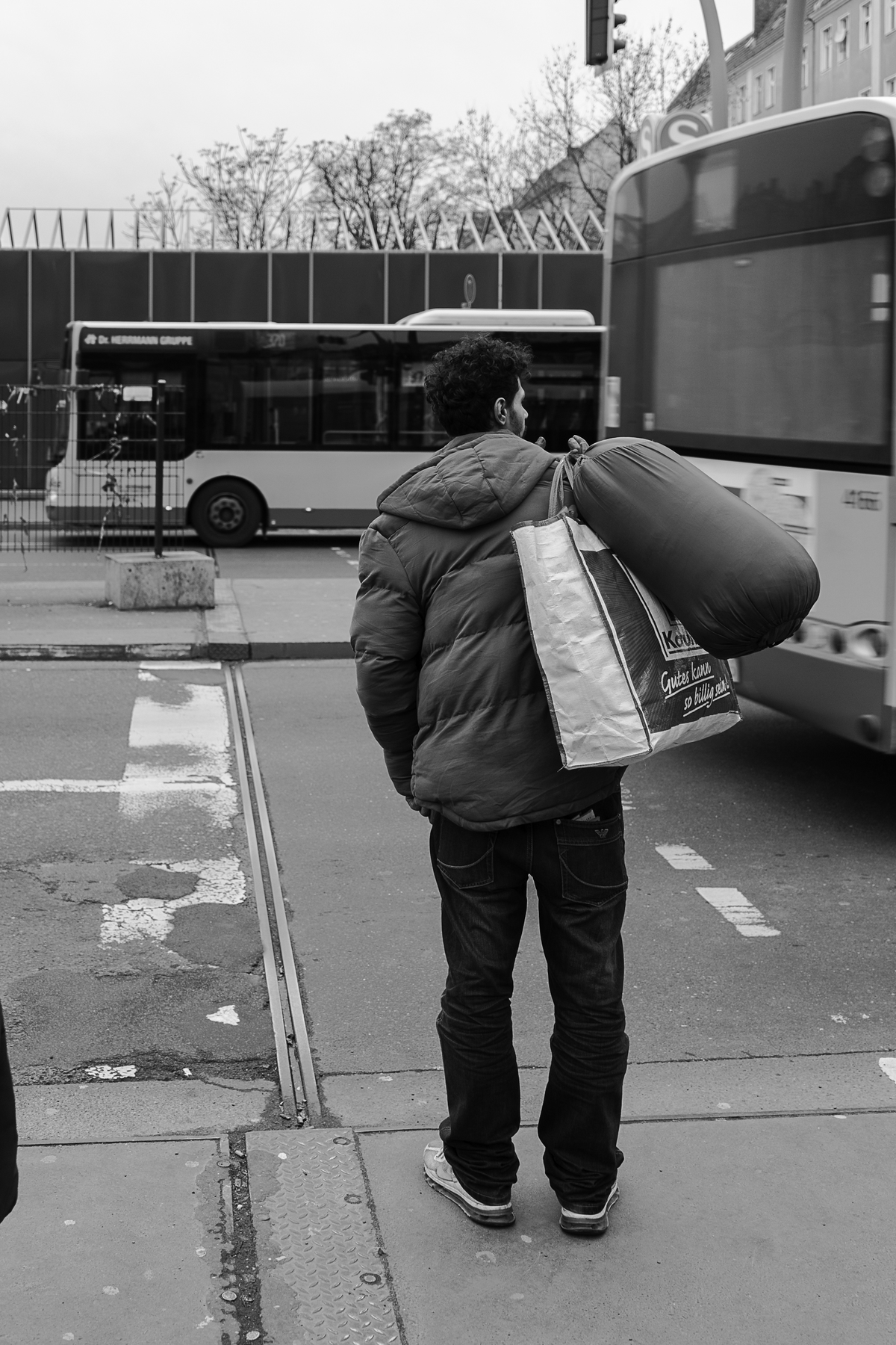 Eine Person in einer Daunenjacke steht an einer Straßenecke und trägt eine große Tasche und ein Bündel, im Hintergrund sind Stadtbusse zu sehen. (KI generierte Beschreibung)