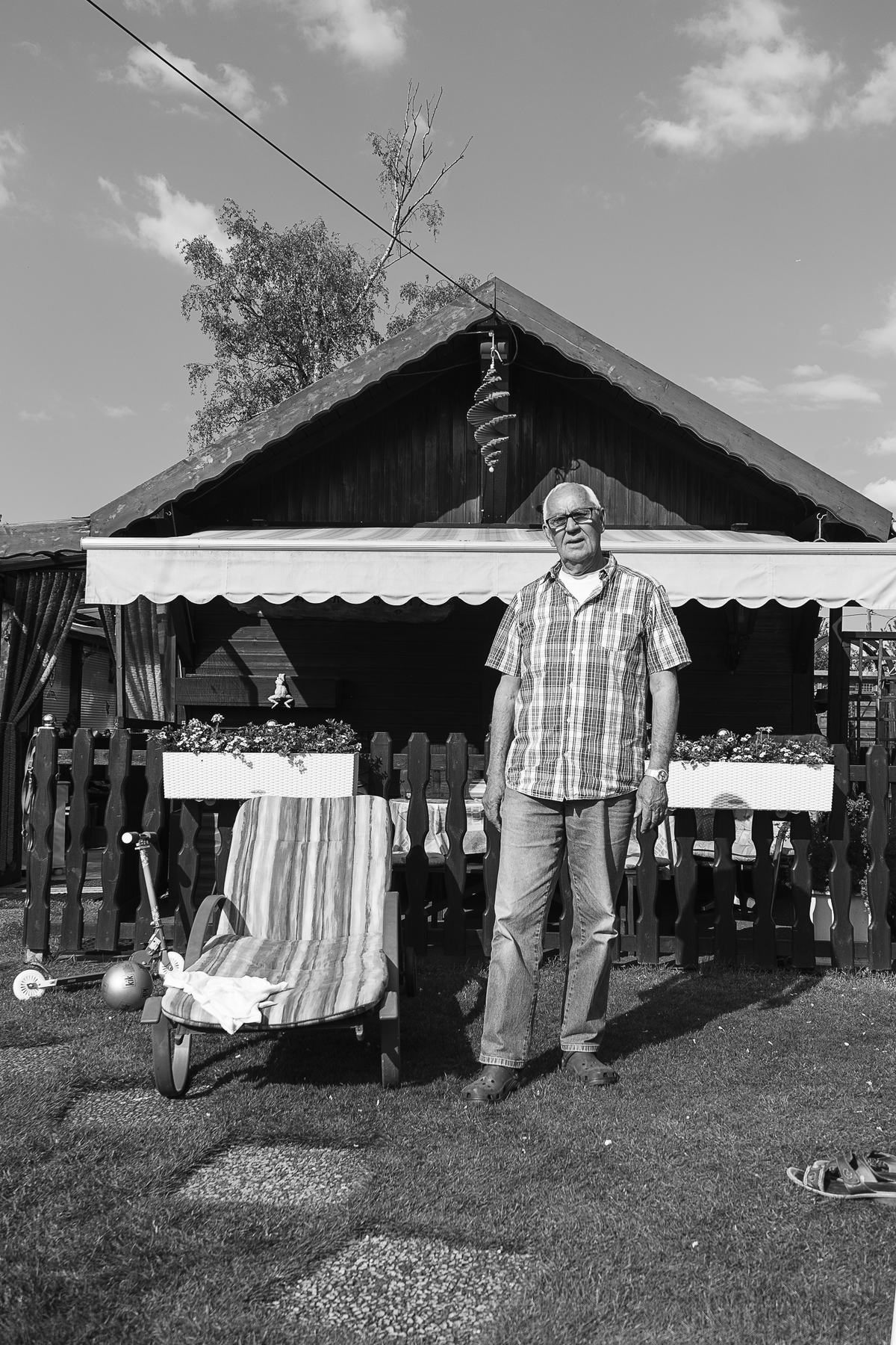 Ein älterer Mann steht in einem Garten vor einem kleinen Holzhaus, neben dem ein Liegestuhl steht. (KI generierte Beschreibung)