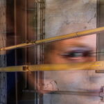 Unscharfes Bild von überlappenden Rohren und einem teilweise sichtbaren Gesicht, mit einer strukturierten, rissigen Wand im Hintergrund. (KI generierte Beschreibung)