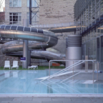 Ein Freibad mit klarem Wasser, einer Metallbadeleiter, mehreren weißen Liegestühlen und einer großen, gewundenen, überdachten Wasserrutsche, die an ein modernes Glasgebäude angebaut ist. Im Hintergrund sind Bäume und Backsteinmauern zu sehen. (KI generierte Beschreibung)