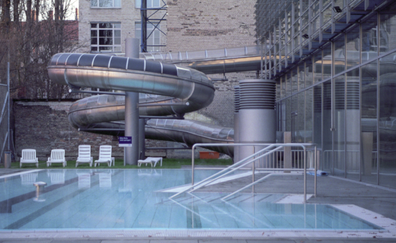 Ein Freibad mit einer großen Metallröhrenrutsche und weißen Liegestühlen auf der Terrasse. (KI generierte Beschreibung)