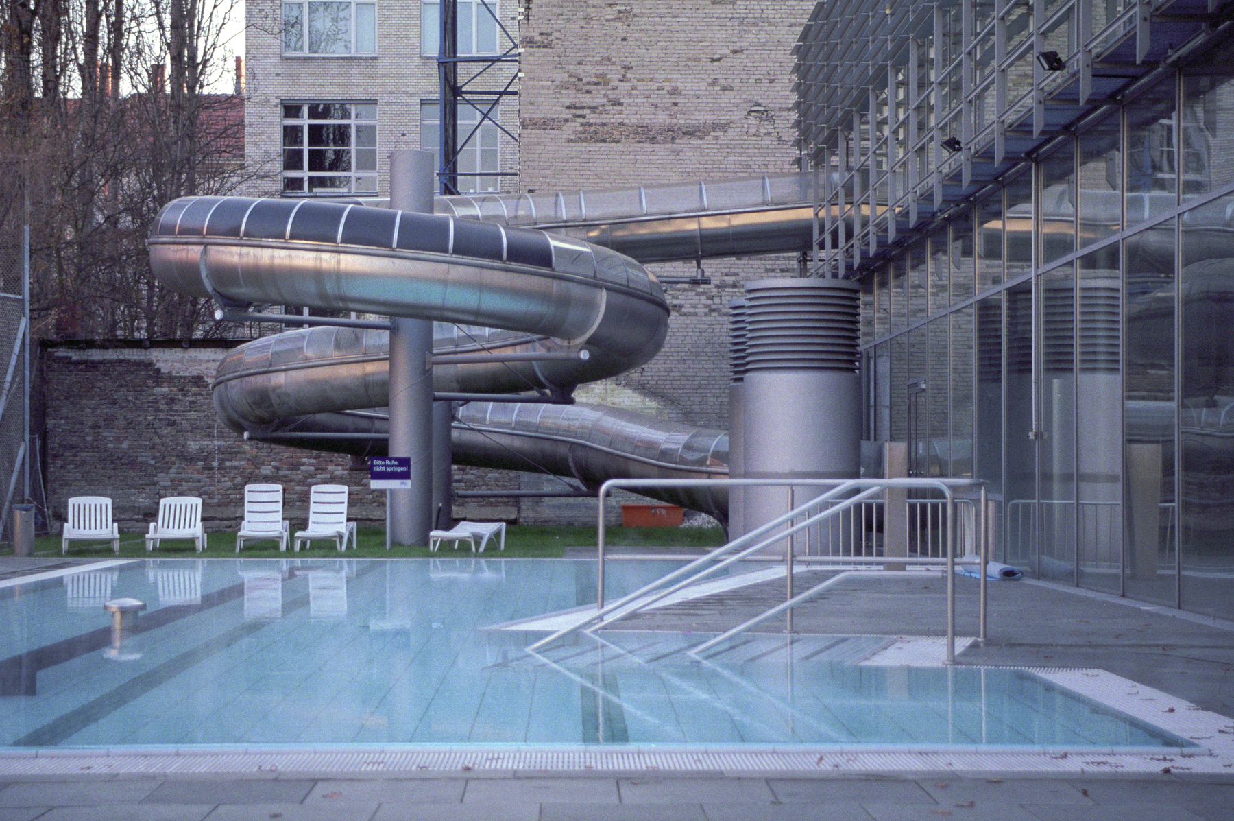 Ein Freibad mit einer großen Metallröhrenrutsche und weißen Liegestühlen auf der Terrasse. (KI generierte Beschreibung)