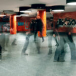 Verschwommene Gestalten gehen durch einen unterirdischen Gang mit orange gekachelten Säulen und Graffiti-bedeckten Wänden. (KI generierte Beschreibung)