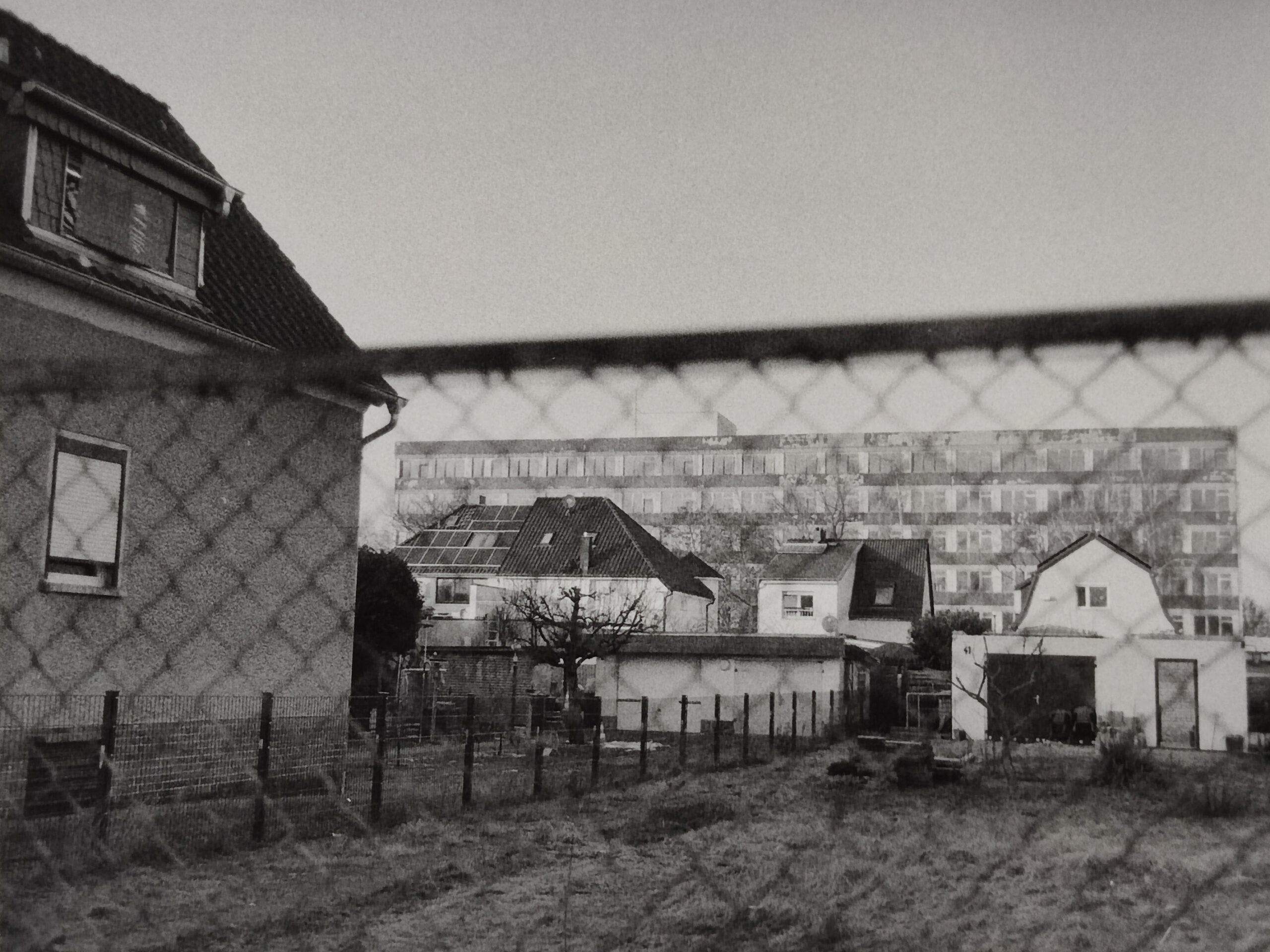 Schwarz-Weiß-Foto, das einen umzäunten Hof mit mehreren Häusern zeigt; im Hintergrund ist durch den Maschendrahtzaun ein großes Mehrfamilienhaus zu sehen. (KI generierte Beschreibung)