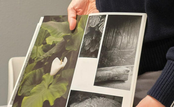Eine Person hält ein Buch offen, in dem vier Fotos zu sehen sind: eine weiße Blume, Dschungelpflanzen, ein Wald und ein auf Holz ruhender Krokodilskopf. (KI generierte Beschreibung)