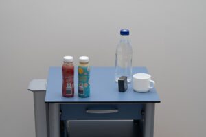 Ein Tisch mit zwei Smoothie-Flaschen, einer durchsichtigen Wasserflasche, einem weißen Becher und einem metallischen Gegenstand vor einem neutralen Hintergrund.