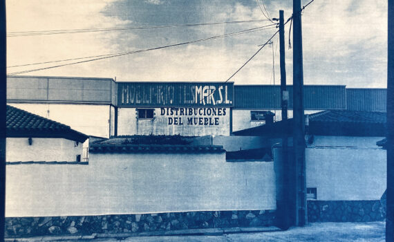 Ein altes Foto eines Möbelvertriebsgebäudes mit Schildern mit der Aufschrift „Distribuciones del Mueble“ und teilweiser Sichtbarkeit von „Muebles y Muebles S.L.“, umgeben von einer niedrigen Mauer. (KI generierte Beschreibung)