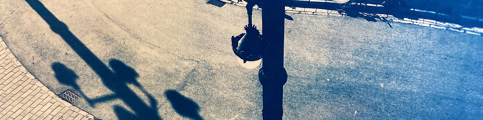 Draufsicht auf eine Straßenszene, die zwei Menschen zeigt, die auf einem geschwungenen Weg neben einem Bürgersteig gehen, im Schatten einer großen Straßenlaterne. Das Bild hat einen blauen Farbton. (KI generierte Beschreibung)
