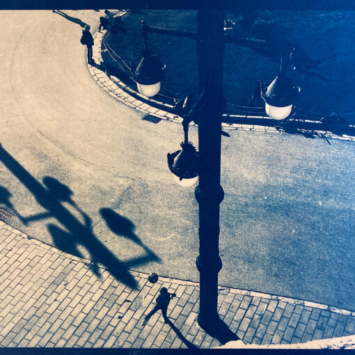Draufsicht auf eine Straßenszene, die zwei Menschen zeigt, die auf einem geschwungenen Weg neben einem Bürgersteig gehen, im Schatten einer großen Straßenlaterne. Das Bild hat einen blauen Farbton. (KI generierte Beschreibung)