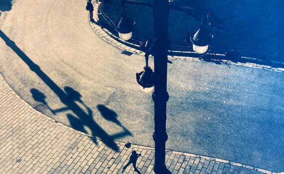 Draufsicht auf eine Straßenszene, die zwei Menschen zeigt, die auf einem geschwungenen Weg neben einem Bürgersteig gehen, im Schatten einer großen Straßenlaterne. Das Bild hat einen blauen Farbton. (KI generierte Beschreibung)