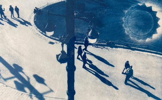 Cyanotypie-Bild von Menschen, die auf einem sonnenbeschienenen Platz spazieren gehen und lange Schatten werfen. Auf dem Boden neben einem Laternenpfahl ist ein kreisförmiges Muster zu sehen. (KI generierte Beschreibung)
