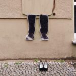 Pappaufsteller von Beinen an einer Wand über einem Gehweg, davor auf dem Boden ein Paar schwarze High Heels. (KI generierte Beschreibung)