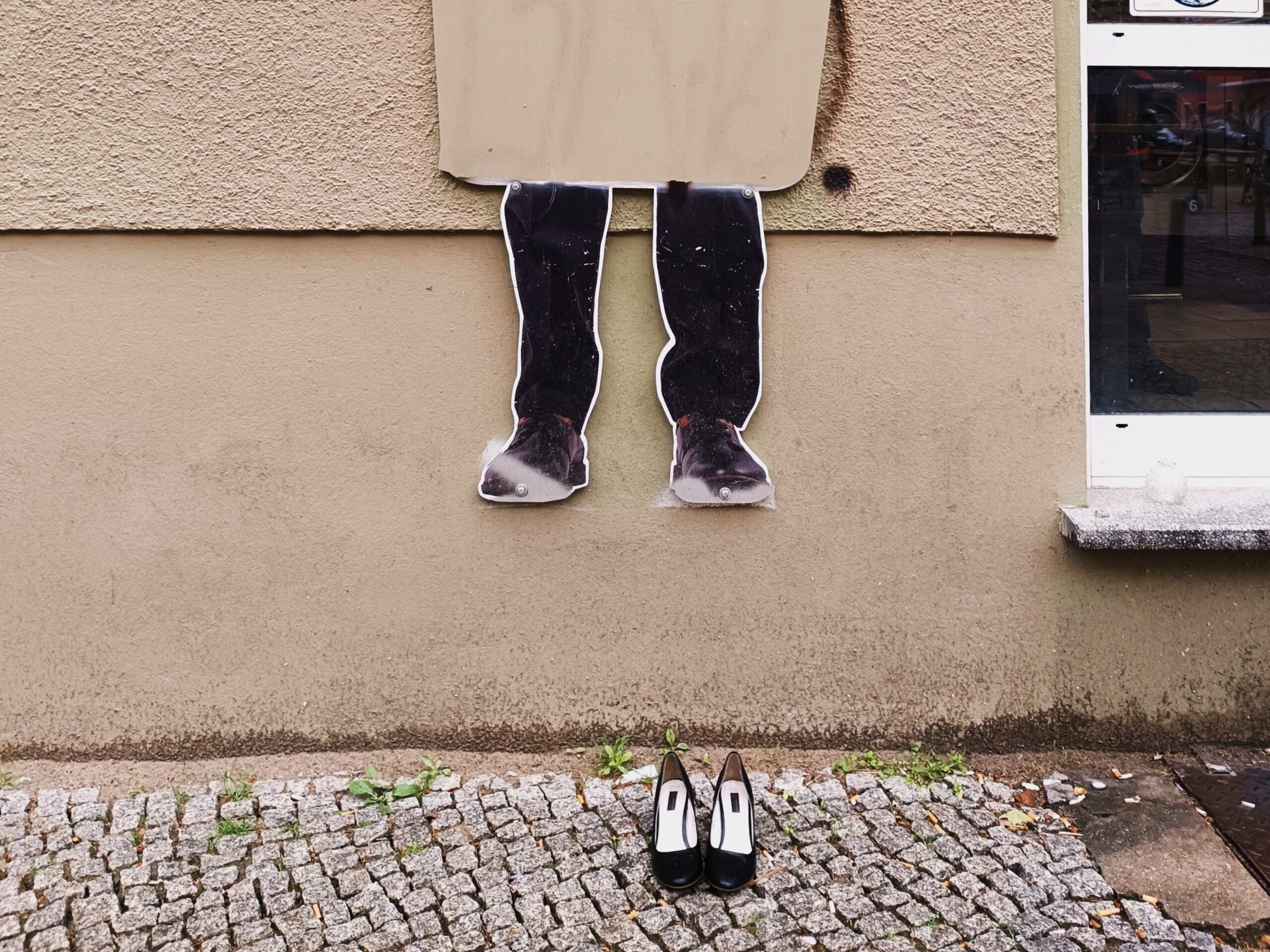 Pappaufsteller von Beinen an einer Wand über einem Gehweg, davor auf dem Boden ein Paar schwarze High Heels. (KI generierte Beschreibung)