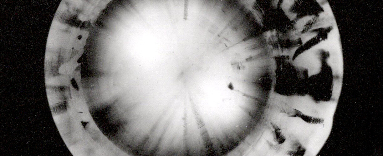 Ein kreisförmiges, abstraktes Schwarz-Weiß-Muster, das einer sich drehenden Scheibe oder Linse ähnelt und sich vor einem schwarzen Hintergrund abhebt. (KI generierte Beschreibung)
