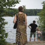 Eine Frau in einem gemusterten Kleid steht an einem See, während ein Mann und ein Kind in Badeanzügen am Ufer stehen. Bäume umgeben die Szene. (KI generierte Beschreibung)
