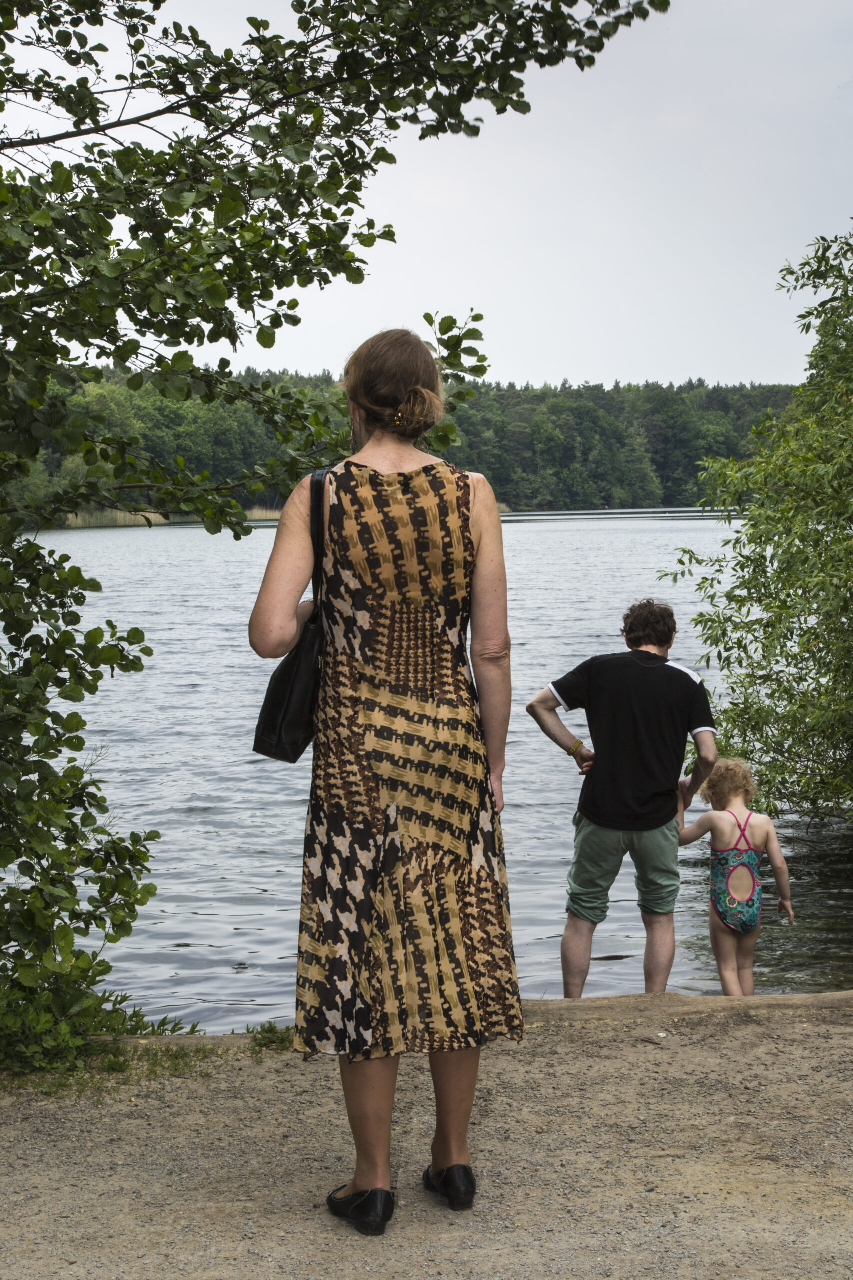 Eine Frau in einem gemusterten Kleid steht an einem See, während ein Mann und ein Kind in Badeanzügen am Ufer stehen. Bäume umgeben die Szene. (KI generierte Beschreibung)