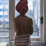Eine Person, die sich ein Handtuch um Kopf und Körper gewickelt hat, steht in einem Raum vor einem Fenster und schaut nach draußen. (KI generierte Beschreibung)