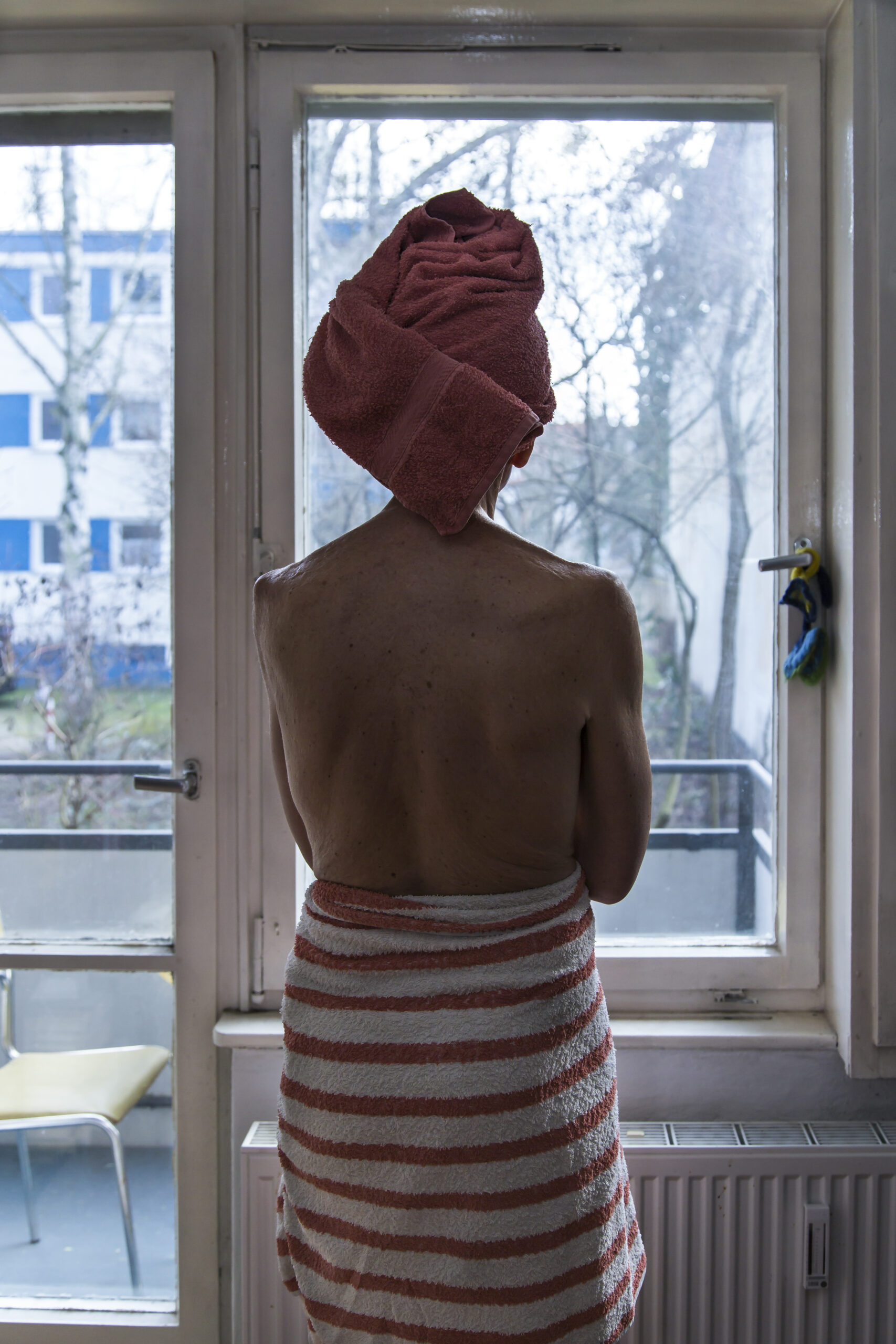Eine Person, die sich ein Handtuch um Kopf und Körper gewickelt hat, steht in einem Raum vor einem Fenster und schaut nach draußen. (KI generierte Beschreibung)
