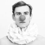 Schwarz-Weiß-Foto einer Person, die einen großen Rüschenkragen und eine runde rote Clownsnase trägt und vor einem schlichten Hintergrund direkt in die Kamera blickt. (KI generierte Beschreibung)