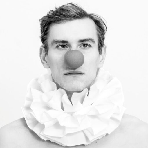 Schwarz-Weiß-Foto einer Person, die einen großen Rüschenkragen und eine runde rote Clownsnase trägt und vor einem schlichten Hintergrund direkt in die Kamera blickt. (KI generierte Beschreibung)