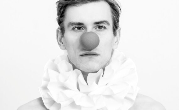 Schwarz-Weiß-Foto einer Person, die einen großen Rüschenkragen und eine runde rote Clownsnase trägt und vor einem schlichten Hintergrund direkt in die Kamera blickt. (KI generierte Beschreibung)