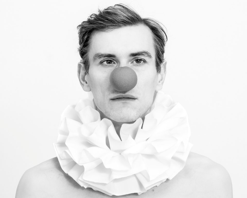 Schwarz-Weiß-Foto einer Person, die einen großen Rüschenkragen und eine runde rote Clownsnase trägt und vor einem schlichten Hintergrund direkt in die Kamera blickt. (KI generierte Beschreibung)