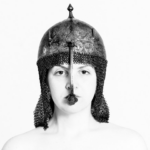 Eine Person, die einen Helm im mittelalterlichen Stil mit Kettenhemd trägt und vor einem schlichten weißen Hintergrund nach vorne blickt. (KI generierte Beschreibung)