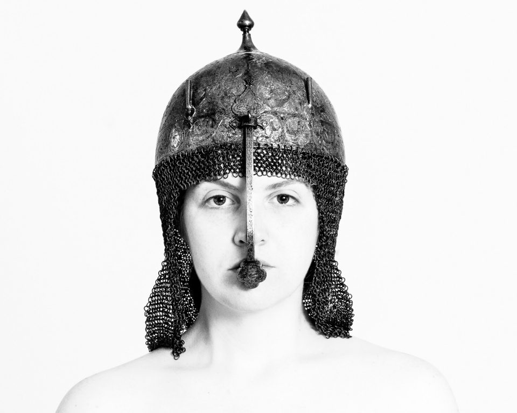 Eine Person, die einen Helm im mittelalterlichen Stil mit Kettenhemd trägt und vor einem schlichten weißen Hintergrund nach vorne blickt. (KI generierte Beschreibung)
