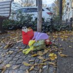 Eine McDonald's Happy Meal-Schachtel, eine Plastiktüte und eine kleine Elfenpuppe liegen auf einem mit Laub bedeckten Gehweg neben einer Bank und einem Baum. (KI generierte Beschreibung)