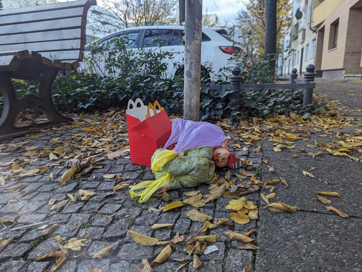 Eine McDonald's Happy Meal-Schachtel, eine Plastiktüte und eine kleine Elfenpuppe liegen auf einem mit Laub bedeckten Gehweg neben einer Bank und einem Baum. (KI generierte Beschreibung)