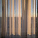 Vor einem Fenster hängen beigefarbene, durchsichtige Vorhänge, die weiches natürliches Licht hereinlassen und sanfte Schatten werfen, mit einem undeutlichen Blick nach draußen im Hintergrund. (KI generierte Beschreibung)