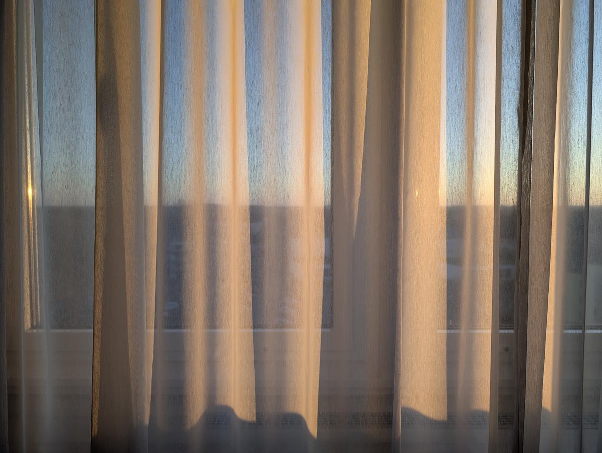 Vor einem Fenster hängen beigefarbene, durchsichtige Vorhänge, die weiches natürliches Licht hereinlassen und sanfte Schatten werfen, mit einem undeutlichen Blick nach draußen im Hintergrund. (KI generierte Beschreibung)