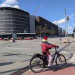 Eine Person, die eine rote Jacke und einen gelben Schal trägt, fährt mit dem Fahrrad über eine Straße in der Stadt; im Hintergrund sind ein Bus und moderne Gebäude bei teilweise bewölktem Himmel zu sehen. (KI generierte Beschreibung)