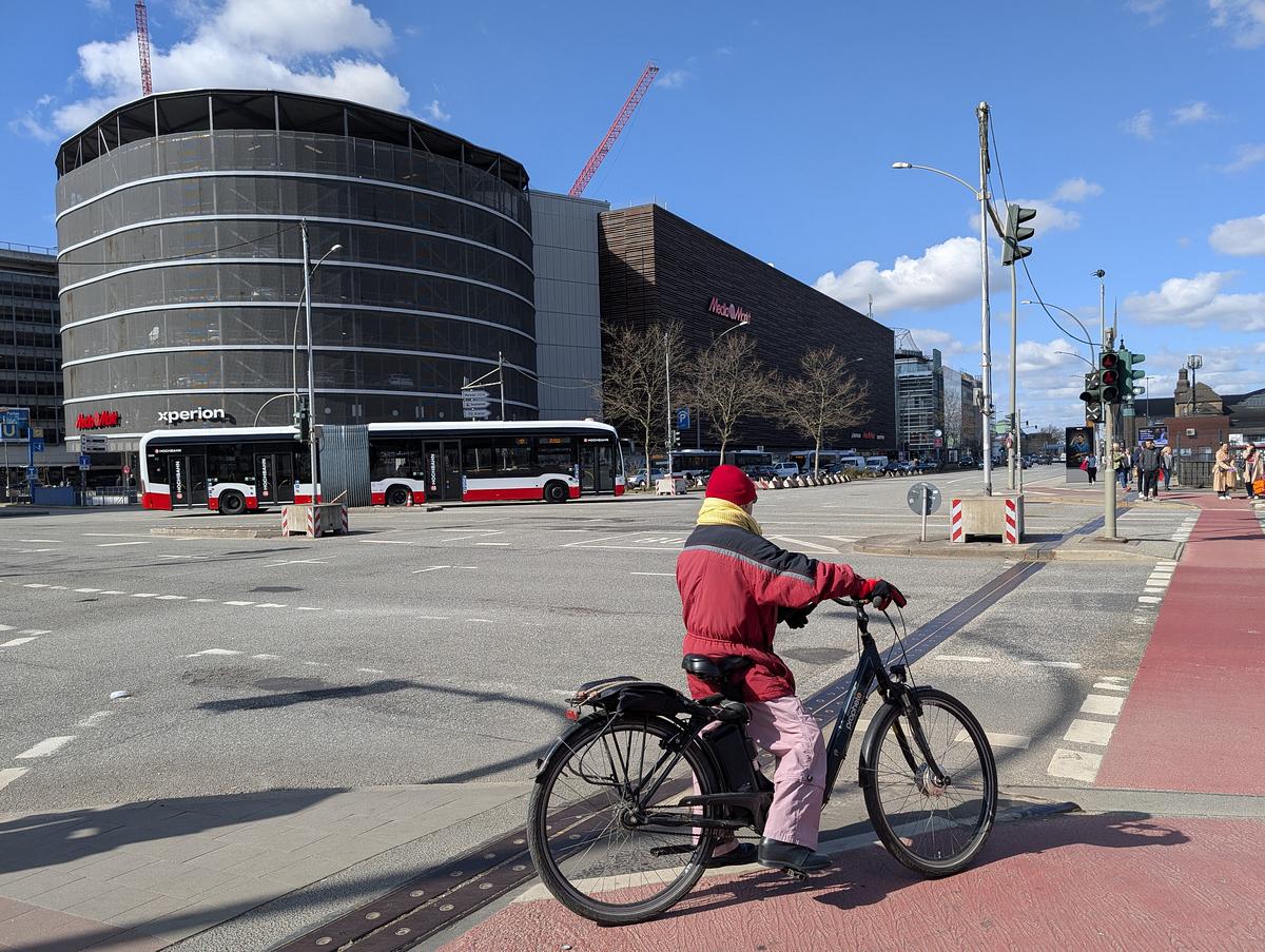 Eine Person, die eine rote Jacke und einen gelben Schal trägt, fährt mit dem Fahrrad über eine Straße in der Stadt; im Hintergrund sind ein Bus und moderne Gebäude bei teilweise bewölktem Himmel zu sehen. (KI generierte Beschreibung)