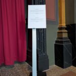 Ein Metallständer mit einem aufgedruckten Hinweis steht neben einer verzierten Tür mit roten Vorhängen und dekorativen schwarz-goldenen Verzierungen. (KI generierte Beschreibung)