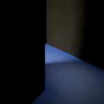Eine dunkle Wand und eine Tür geben teilweise den Blick auf einen Raum mit blauem Boden frei, der von einer unsichtbaren Lichtquelle beleuchtet wird. (KI generierte Beschreibung)