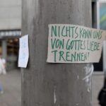 Auf einem handgeschriebenen Pappschild an einem Mast steht "NICHTS KANN UNS VON GOTTES LIEBE TRENNEN!" in deutscher Sprache; im Hintergrund sind eine Person und Gebäude zu sehen. (KI generierte Beschreibung)