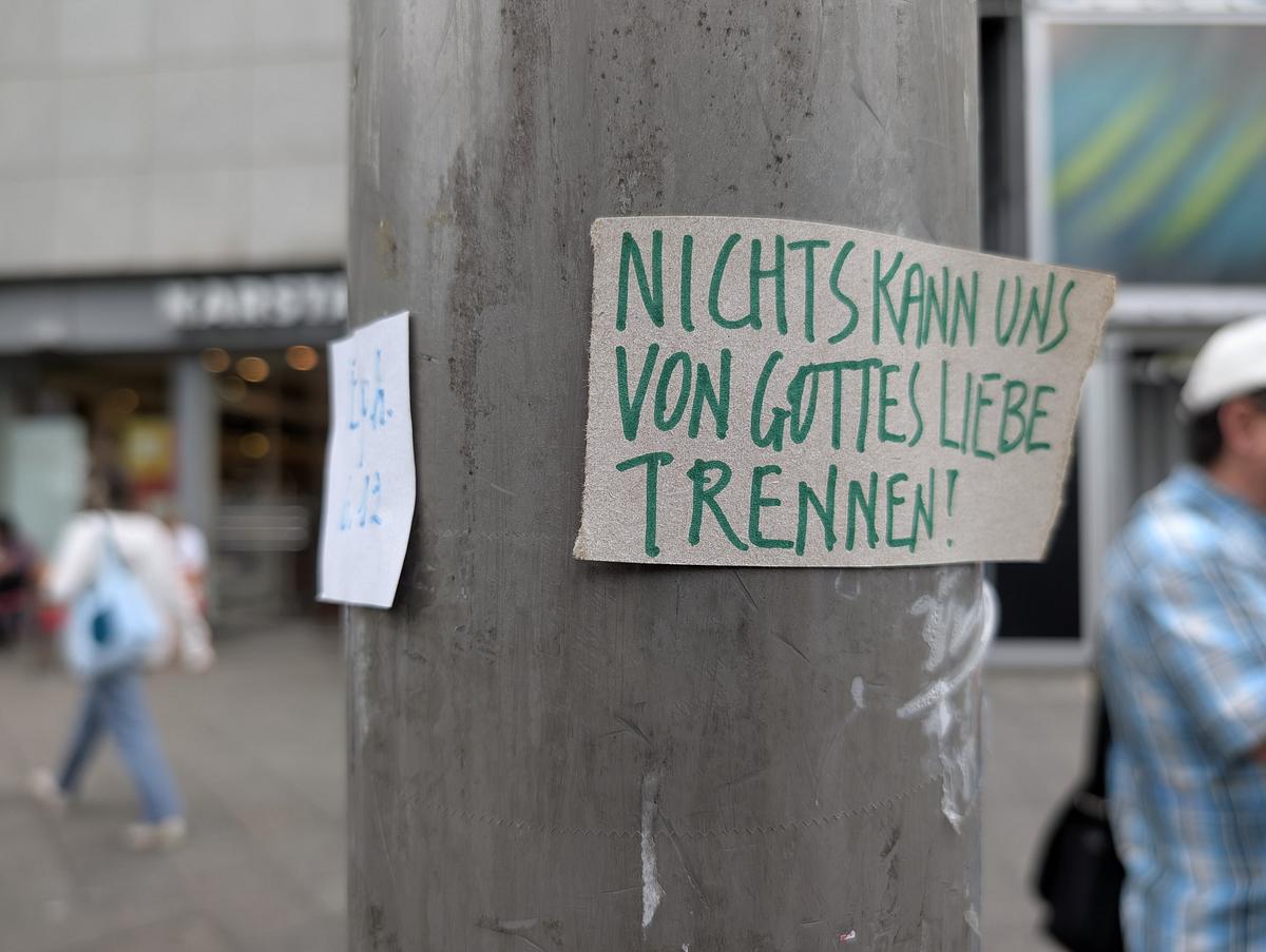 Auf einem handgeschriebenen Pappschild an einem Mast steht "NICHTS KANN UNS VON GOTTES LIEBE TRENNEN!" in deutscher Sprache; im Hintergrund sind eine Person und Gebäude zu sehen. (KI generierte Beschreibung)