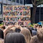 Eine Menschenmenge hält ein buntes Schild mit der Aufschrift "Es gibt so viel Menschlichkeit, Liebe und Schönheit, für die es sich zu kämpfen lohnt - Gina Martin" bei einer Versammlung im Freien hoch. (KI generierte Beschreibung)