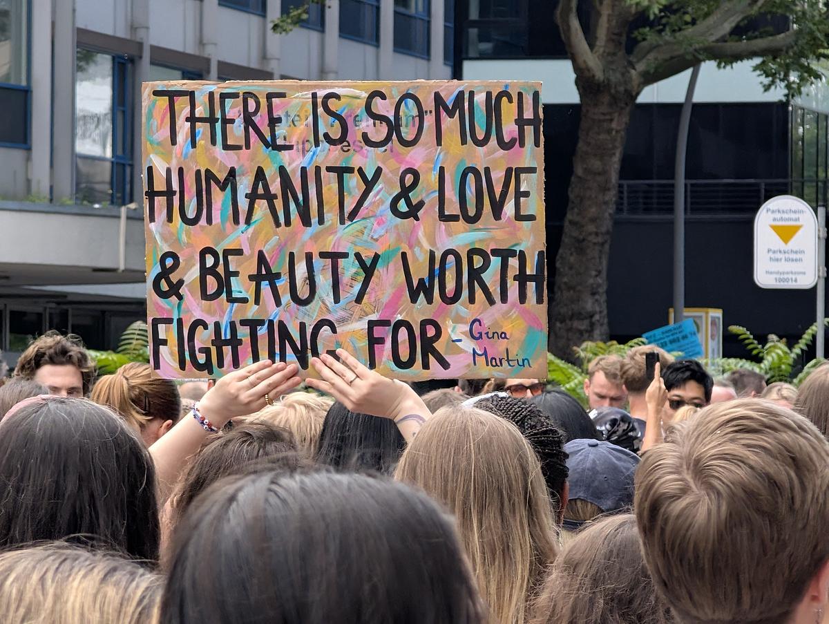 Eine Menschenmenge hält ein buntes Schild mit der Aufschrift "Es gibt so viel Menschlichkeit, Liebe und Schönheit, für die es sich zu kämpfen lohnt - Gina Martin" bei einer Versammlung im Freien hoch. (KI generierte Beschreibung)