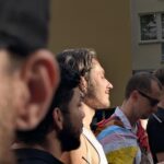 Eine Gruppe von Menschen steht im Freien, eine Person ist in eine Progress Pride-Flagge gehüllt, und das Sonnenlicht beleuchtet einen Mann in der Mitte. (KI generierte Beschreibung)