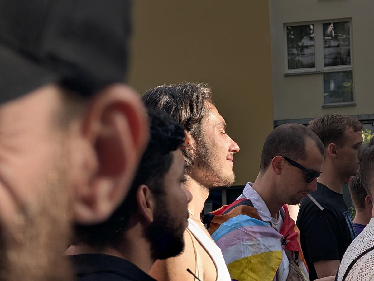 Eine Gruppe von Menschen steht im Freien, eine Person ist in eine Progress Pride-Flagge gehüllt, und das Sonnenlicht beleuchtet einen Mann in der Mitte. (KI generierte Beschreibung)