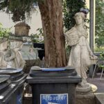 Zwei Steinstatuen stehen in der Nähe von schwarzen Recyclingbehältern mit der Aufschrift "Altpapier" in einer Außenanlage mit Pflanzen, Bäumen und Sitzgelegenheiten im Hintergrund. (KI generierte Beschreibung)