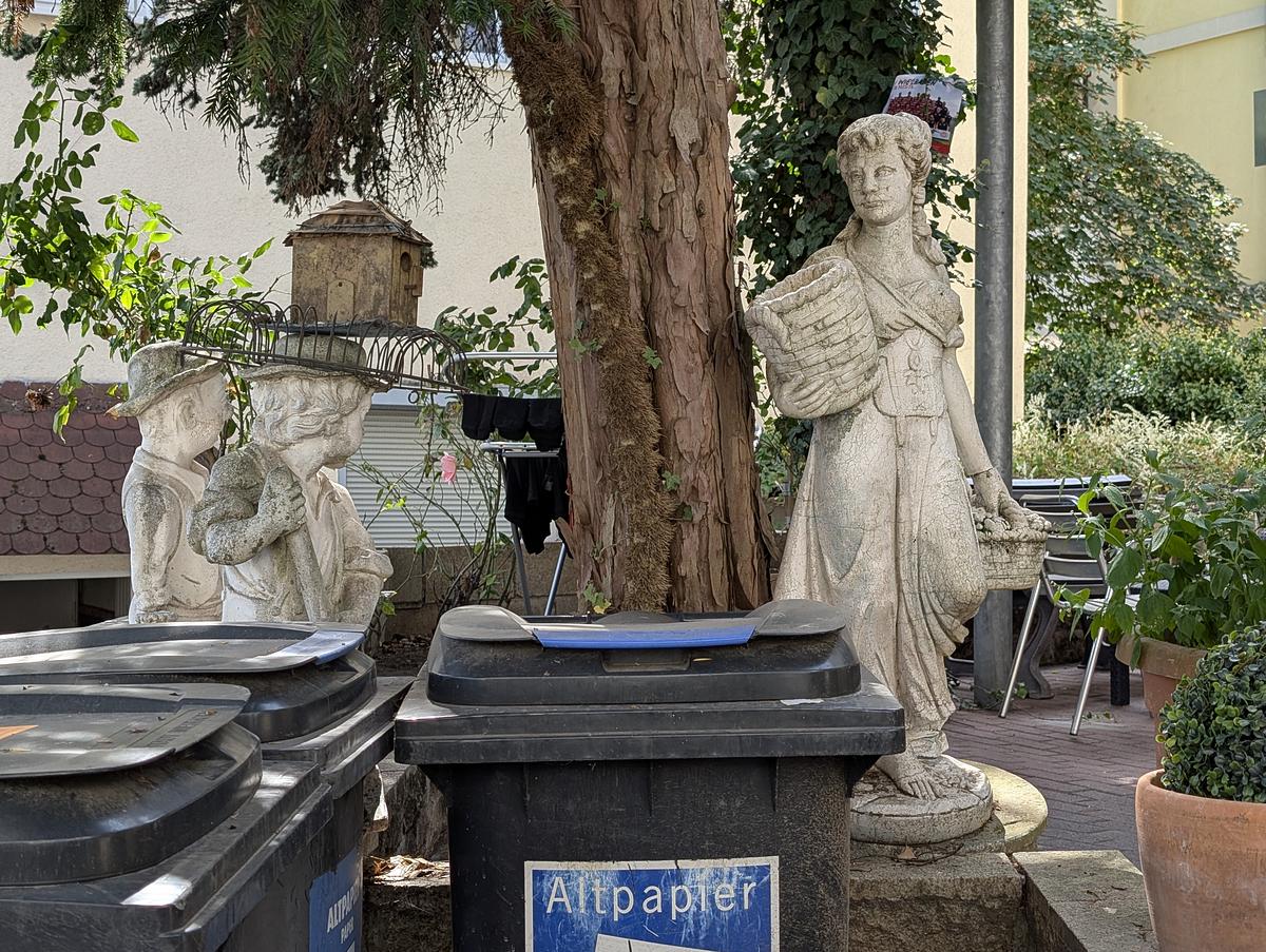 Zwei Steinstatuen stehen in der Nähe von schwarzen Recyclingbehältern mit der Aufschrift "Altpapier" in einer Außenanlage mit Pflanzen, Bäumen und Sitzgelegenheiten im Hintergrund. (KI generierte Beschreibung)